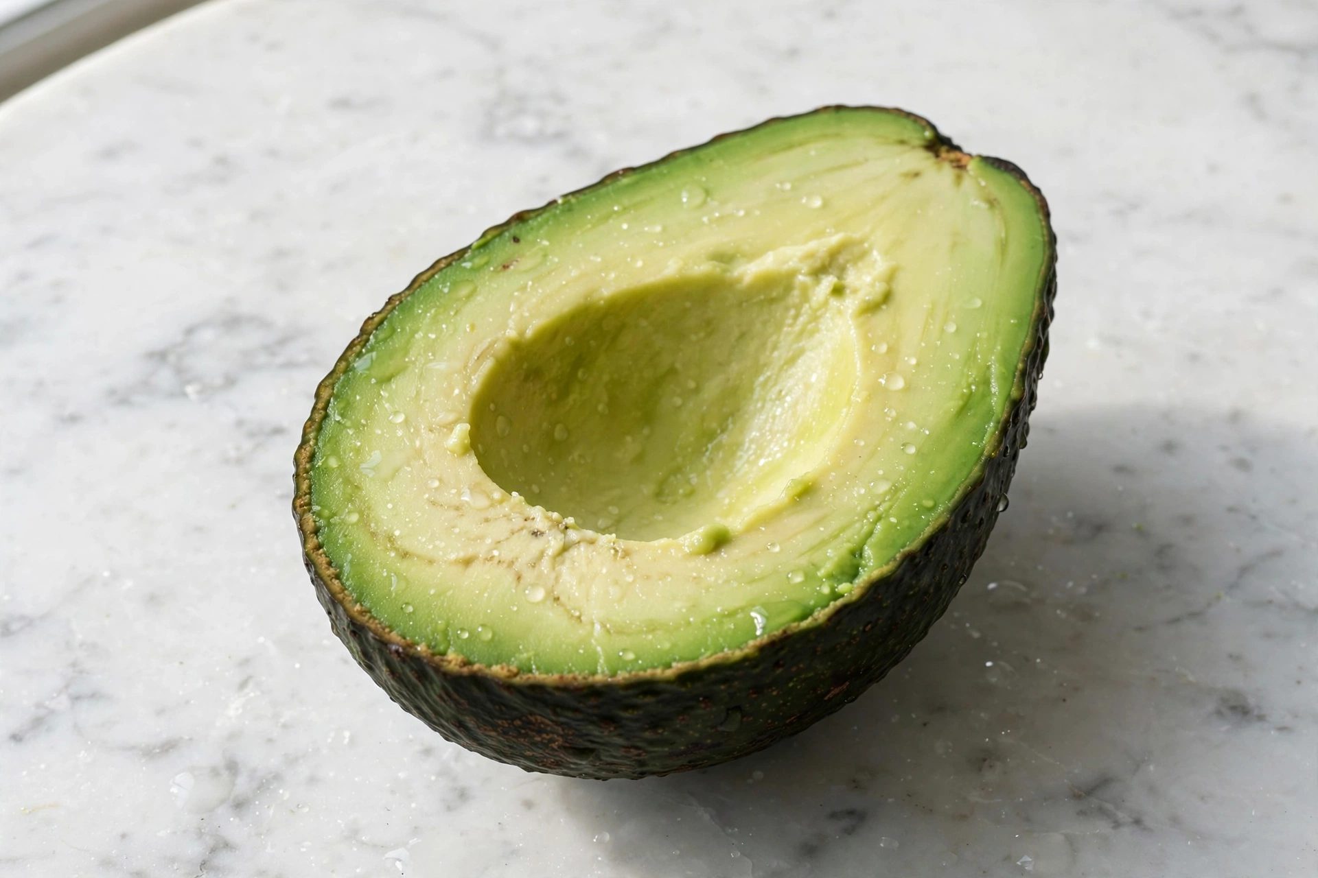 Mezzo avocado maturo con la cavità del nocciolo vuota, appoggiato su un piano di marmo bianco